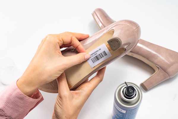 Een hand houdt een schoen met een prijskaartje, terwijl er een spuitbus naast staat. De schoen is een beige hak.