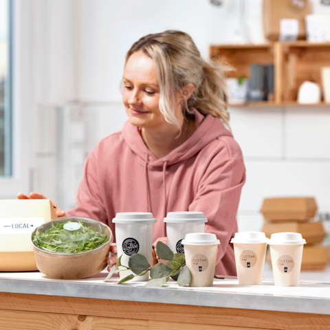 Een vrouw in een roze hoodie bereidt gezonde maaltijden voor, waaronder een kom met salade en verschillende koffiekopjes op een tafel.