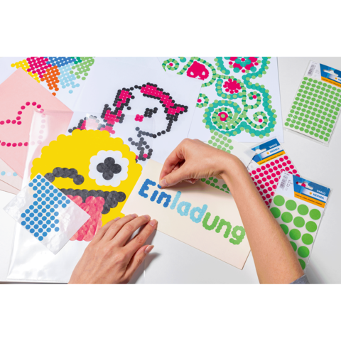 Eine Hand platziert bunte Punkte auf ein Einladungskarte, um ein kreatives Design zu erstellen. Verschiedene Bastelmaterialien sind sichtbar.
