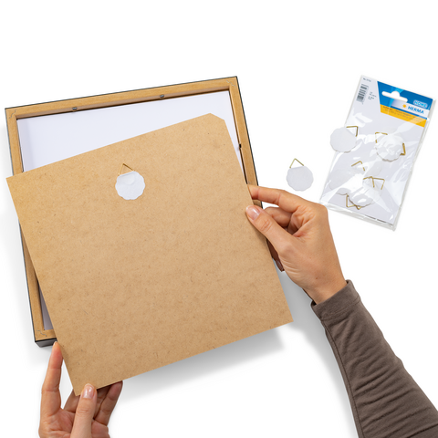 Eine Person hält eine Rückwand aus Pappe mit einem Loch, während eine Bilderrahmenverpackung und Aufhänger im Hintergrund sichtbar sind.
