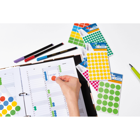 Ein geöffneter Planer mit bunten Aufklebern und Stiften. Eine Hand platziert einen roten Aufkleber auf einem Datum im Planer.