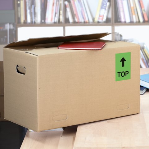 Ein Karton mit dem Aufkleber "FRAGILE", daneben ein graues Kuvert mit dem Aufkleber "URGENT!" und eine Schere auf einem Holztisch.