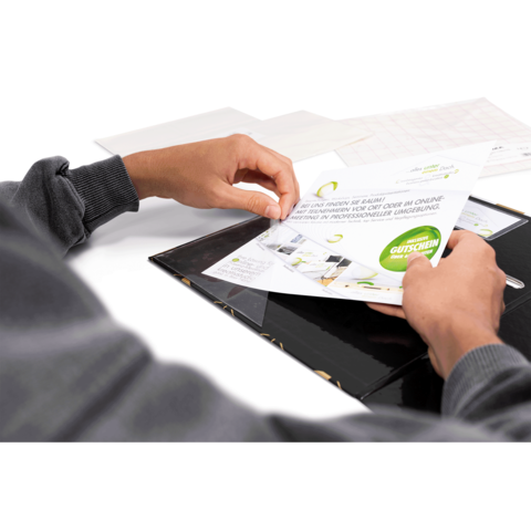 Eine Hand hält einen Gutschein, während sie ihn aus einer schwarzen Mappe zieht. Im Hintergrund sind weitere Dokumente sichtbar.