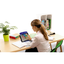 Eine Frau mit langen, braunen Haaren sitzt an einem Schreibtisch und nutzt ein Tablet. Im Hintergrund sind Bücher und eine Pflanze sichtbar.