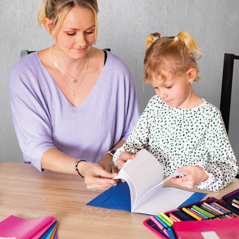Een vrouw en een klein meisje zitten aan een tafel en bladeren door een notitieboek. Kleurrijke stiften liggen verspreid over de tafel.