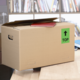 Kartonbox auf einem Tisch, mit einem roten Heft und einem grünen Aufkleber "TOP" auf der Seite. Hintergrund mit Bücherregalen.