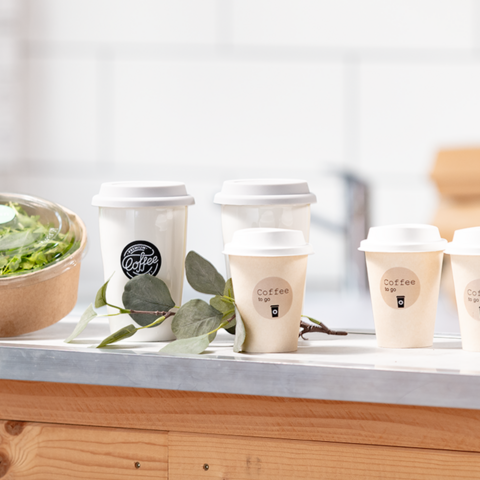 Verschillende koffiebekers staan op een tafel naast een schaal met salade. De bekers zijn voorzien van afhaalopdrukken.