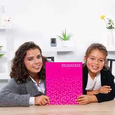 Twee meisjes zitten aan een tafel en houden een roze schrift vast met de opdruk 'GETUIGENISSEN'. Ze glimlachen naar de camera.