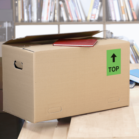 Kartonbox mit einem roten Heft auf der Oberseite, beschriftet mit einem grünen "TOP"-Schild, steht auf einem Tisch vor einem Bücherregal.