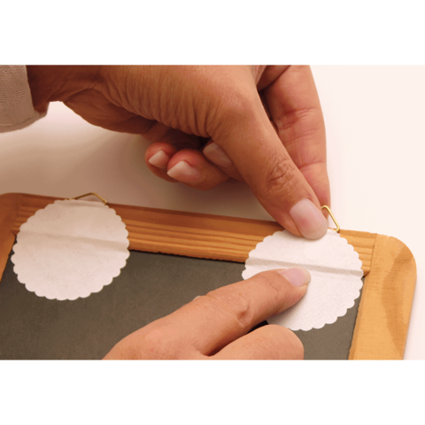 Eine Hand platziert eine runde, weiße Papierschablone auf einer Holztafel, während eine andere Hand einen Haken hält.