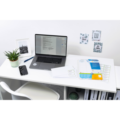 Ein Schreibtisch mit einem Laptop, einem Smartphone, einer Pflanze und verschiedenen Dokumenten. An der Wand hängen Notizen und Bilder.