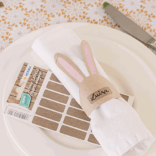 Ein weißer Serviettenring mit Hasenohren und dem Namen "Luisa" auf einer gefalteten Serviette, daneben ein Blatt mit Etiketten.