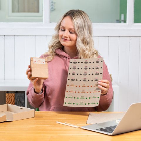Eine Frau mit blonden, lockigen Haaren hält eine kleine Verpackung und ein Blatt mit Aufklebern in einem hellen Raum.