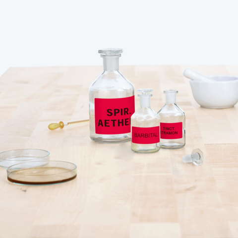 Flaschen mit roten Etiketten, beschriftet mit "SPIR. AETHER", "BARBITAL" und "TINCT. TRAMON", auf einem Holztisch.