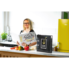 Eine Frau mit Brille hält ein Kochbuch in der Küche, umgeben von frischem Gemüse und einer grünen Pflanze.