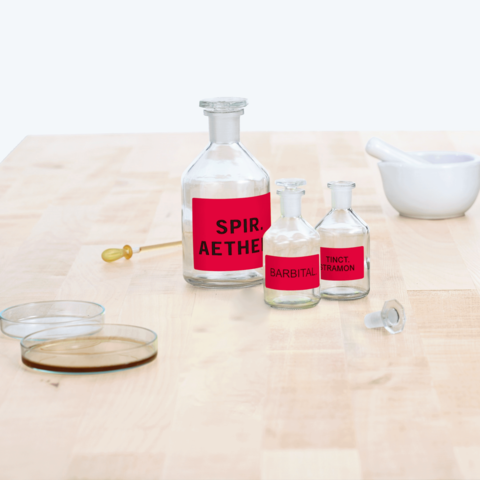 Flaschen mit roten Etiketten, beschriftet mit "Spir. Aether", "Barbital" und "Tinct. Tramon", auf einem Holztisch neben einer Schale.