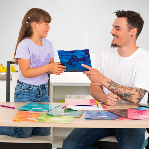 Ein Mann und ein Mädchen sitzen an einem Tisch, während sie farbige Ordner und Papiere betrachten. Der Mann zeigt auf einen Ordner.