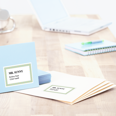 Blau en witte enveloppen met adreslabels liggen op een tafel, naast een laptop en een notitieboek.