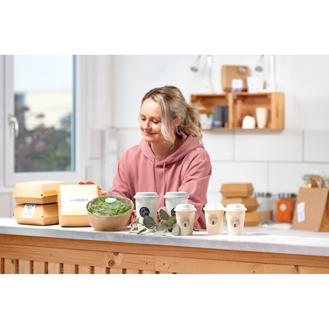 Eine Frau in einem rosa Hoodie steht an einer Küchentheke mit Essensverpackungen und Getränken. Sie lächelt und betrachtet die Schalen.