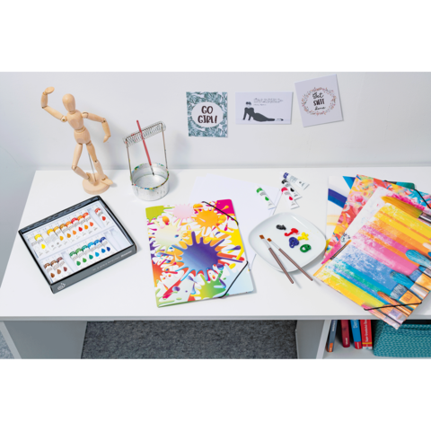 Ein Tisch mit Malutensilien, darunter Aquarellfarben, Pinsel, bunte Papiere und ein Holzmodell. An der Wand hängen motivierende Zitate.