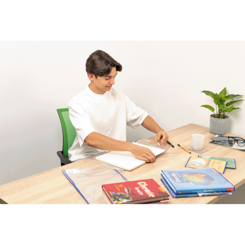 Ein junger Mann sitzt an einem Tisch, blättert in einem Notizbuch und hat Bücher sowie Schreibmaterialien um sich herum. Eine Pflanze steht im Hintergrund.