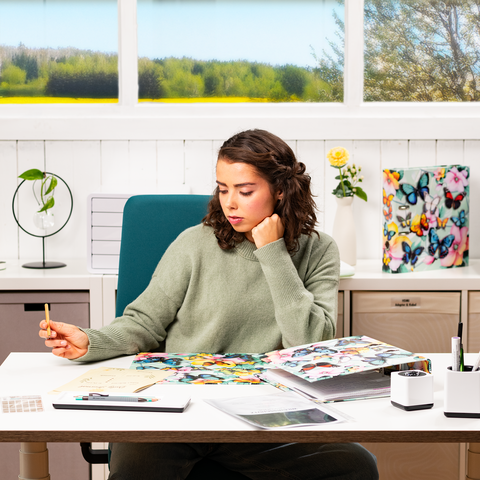 Een jonge vrouw zit aan een bureau, omringd door kleurrijke materialen en aantekeningen, terwijl ze nadenkend een pen vasthoudt.