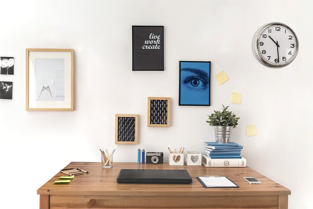 Een bureau met een laptop, decoratieve foto's aan de muur, een plant en een vloerlamp. Een wandklok geeft de tijd aan.
