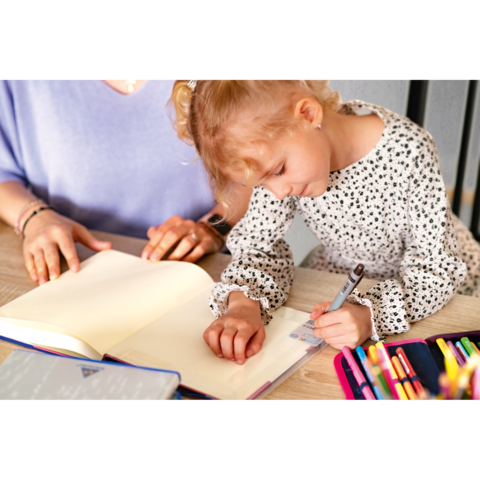 Ein kleines Mädchen schreibt in ein offenes Notizbuch, während eine erwachsene Person daneben sitzt und zuschaut. Bunte Stifte sind sichtbar.