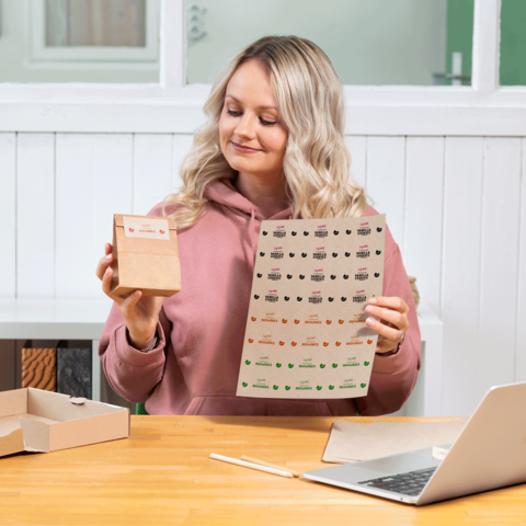 Eine Person mit blonden, lockigen Haaren hält eine kleine Verpackung und ein Blatt mit Aufklebern in einer hellen Umgebung.