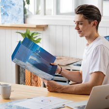 Ein junger Mann sitzt an einem Tisch und betrachtet eine Mappe mit Bildern von Landschaften, während er eine Tasse Kaffee hält.