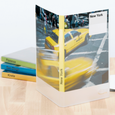 Gelbes Taxi auf einer New Yorker Straße, umgeben von einem transparenten Ordner mit der Aufschrift "New York". Im Hintergrund sind farbige Buchrücken sichtbar.