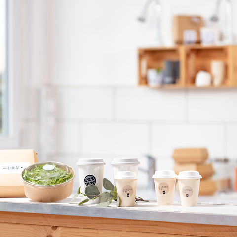 Een houten tafel met een schaal salade, verschillende koffiekopjes en een verpakking, omringd door een lichte, moderne keuken.
