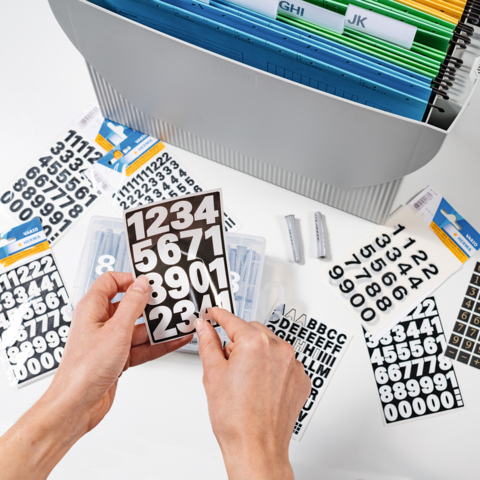 Handen houden een zwart bord met witte cijfers. Op de achtergrond zijn er meer cijfers en letters te zien, evenals een opbergsysteem.