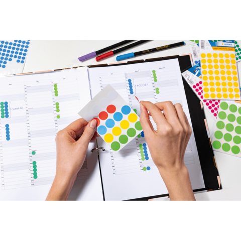 Eine Person hält ein Blatt mit bunten Aufklebern über einem offenen Planer, der mit Notizen und weiteren Aufklebern gefüllt ist.