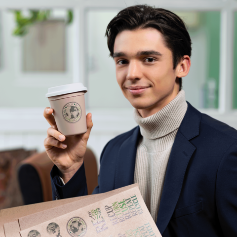 Junger Mann in einem Anzug hält einen Kaffeebecher und lächelt. Im Hintergrund sind unscharfe Möbel und Wände zu sehen.