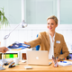 Een vrouw in een kantoor geeft een man een document, terwijl ze aan een bureau met een laptop en kantoormateriaal zit.