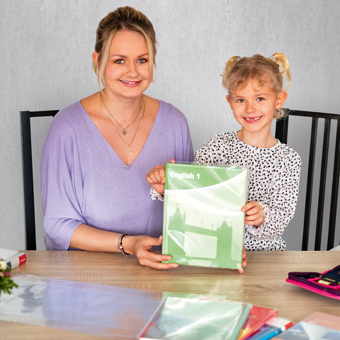 Eine Frau und ein Mädchen sitzen an einem Tisch, das Mädchen hält ein grünes Heft. Auf dem Tisch liegen Schreibwaren und ein Blumenstrauß.