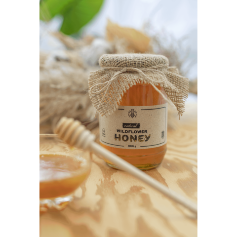 Glas mit Wildblütenhonig, verziert mit einem Jutesack, neben einem kleinen Glas und einem Honiglöffel auf einem Holzuntergrund.