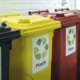 Drei Abfallbehälter in verschiedenen Farben mit Recycling-Symbolen: Gelb für Plastik, Rot für Glas und Schwarz für Restmüll.