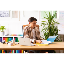 Ein junger Mann sitzt an einem Schreibtisch, arbeitet mit einem Laptop und blättert durch Dokumente in einer Ordnermappe. Pflanzen und farbige Akten sind im Hintergrund.