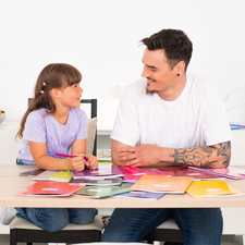 Ein Mann und ein Mädchen sitzen an einem Tisch, umgeben von bunten Farbmusterkarten, und scheinen ein Gespräch zu führen.