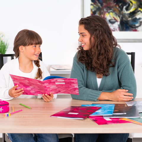 Eine Mutter und ihre Tochter sitzen an einem Tisch, während sie ein Buch betrachten. Auf dem Tisch liegen mehrere farbige Hefte und Schreibmaterialien.