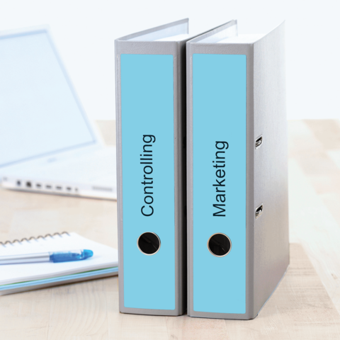Zwei Ordner mit den Beschriftungen "Controlling" und "Marketing" stehen auf einem Tisch, daneben ein Laptop und ein Notizblock.