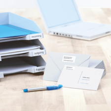 Ordner mit beschrifteten Fächern für eingehende und ausgehende Dokumente, daneben Umschläge und ein blauer Stift auf einem Tisch.