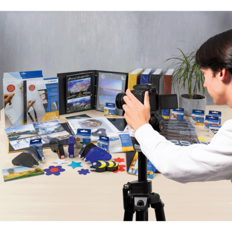 Ein Fotograf steht vor einem Tisch mit verschiedenen Büroartikeln, darunter Ordner, Kleber und Bastelmaterialien, während er eine Kamera hält.