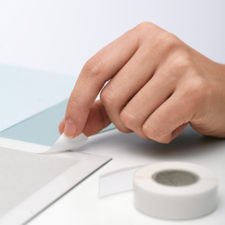 Een hand haalt een stuk tape van een grijze oppervlakte, terwijl er een rol tape naast ligt.