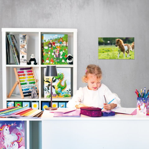 Ein Mädchen sitzt an einem Schreibtisch, schreibt in ein Heft und hat eine Federmappe vor sich. Im Hintergrund sind Regale mit Spielzeug und Kunstmaterialien.