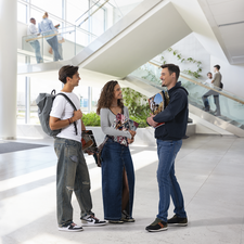 Drei junge Erwachsene stehen in einem modernen Innenraum und unterhalten sich. Sie tragen lässige Kleidung und halten Taschen.