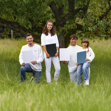 Vier Personen in weißen Oberteilen stehen und sitzen im Gras, halten jeweils Plakate in den Händen. Ein Baum ist im Hintergrund sichtbar.