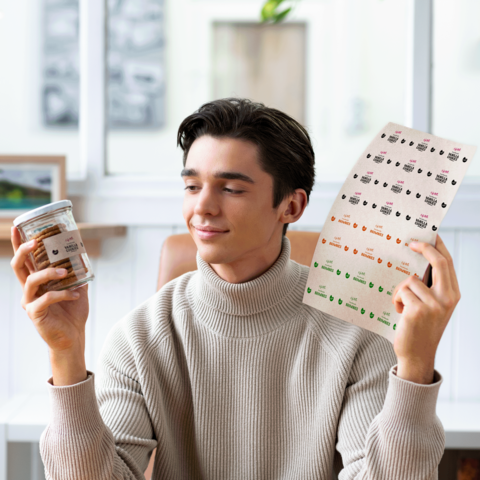 Ein junger Mann hält ein Glas mit Snacks in einer Hand und ein Blatt mit verschiedenen Aufklebern in der anderen Hand, während er lächelt.